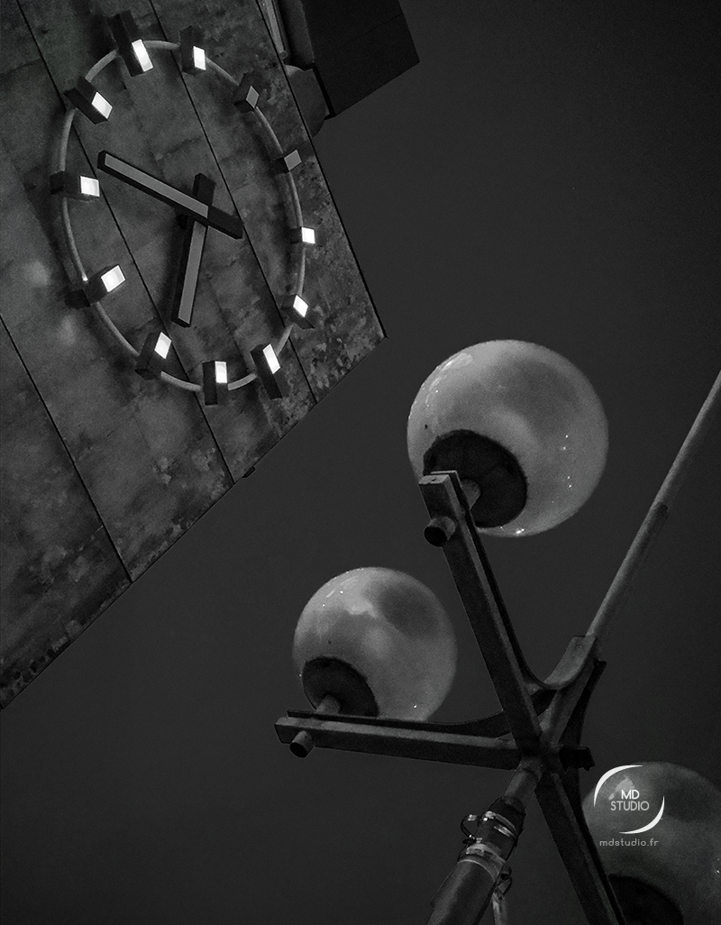 photographie en noir et blanc, lampadaires sphériques au premier plan, horloge ronde en arrière-plan. parvis gare SNCF Sud. Nantes.